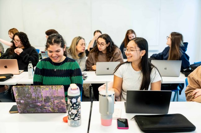 251027-marymount-8258 (1) Students sit together at classroom desks and smile